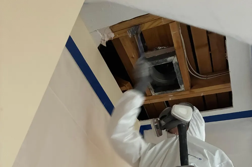 Mold technician in protective suit and respirator opening a ceiling